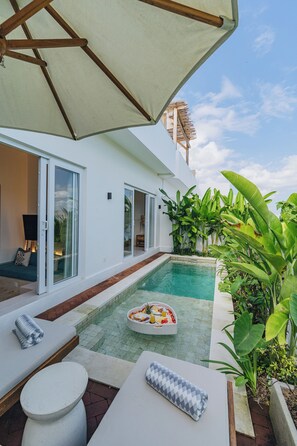 A poolside area with a lounge chair, a small table with a basket of fruit, and a large umbrella providing shade. - Villa Segara Angin (Cemagi)