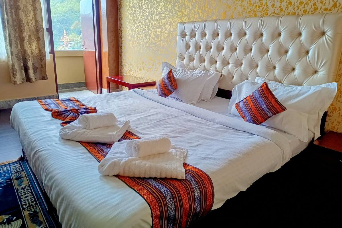 Una cama bien hecha con sábanas blancas y almohadas coloridas, una ventana con cortinas y un papel tapiz estampado.