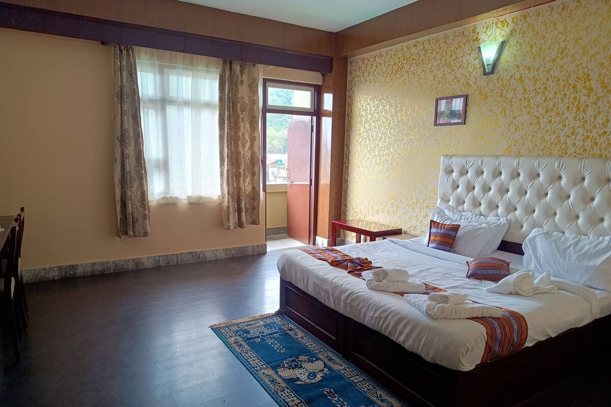 Habitación de hotel con una cama grande, cabecera de madera, alfombra azul estampada y una ventana con cortinas.