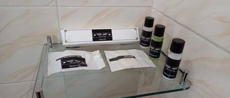 A bathroom with a glass shelf holding a dental kit, shampoo, shower gel, and a bottle of moisturizer.