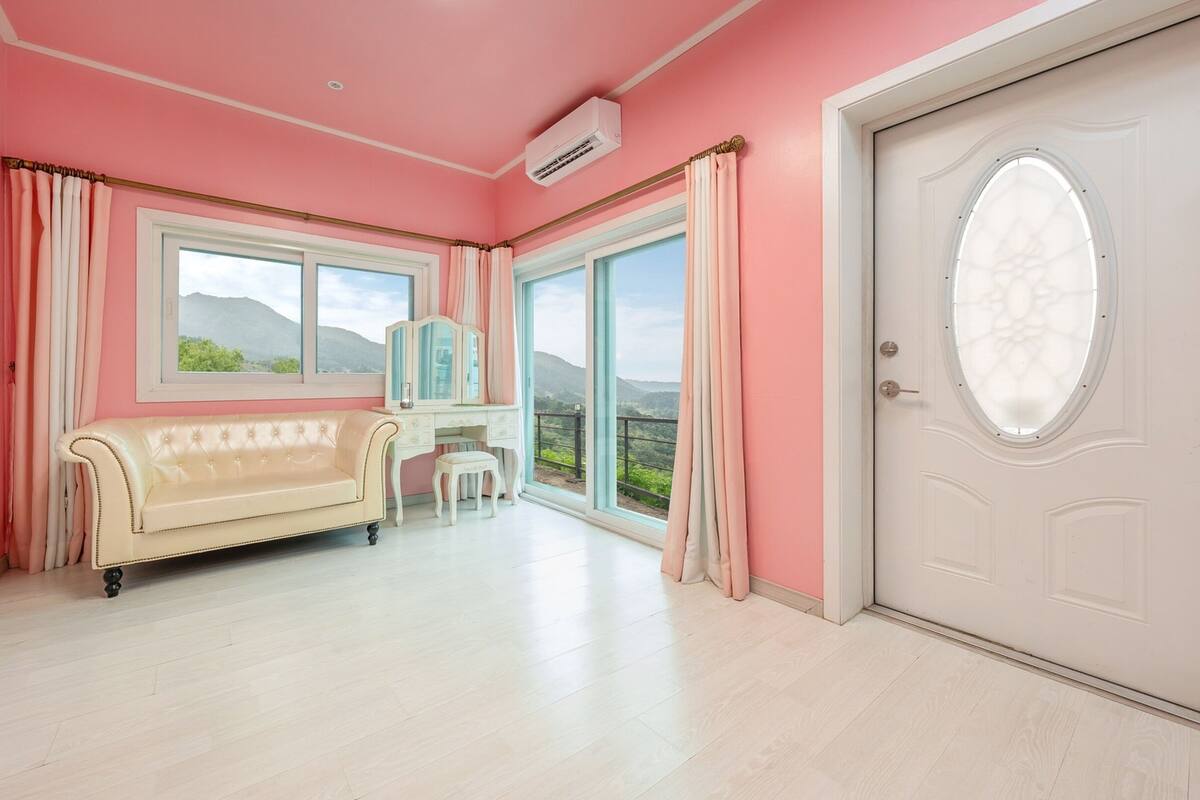 Une pièce avec un mur rose, une porte blanche dotée d’une fenêtre, un canapé beige, une petite table et une vue sur les montagnes.