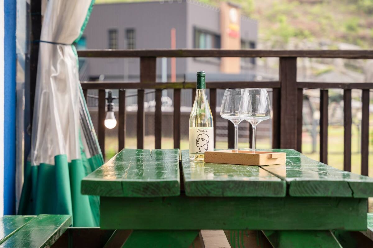 Une table en bois vert, sur laquelle sont posés une bouteille de vin Nadra et deux verres, est placée sur un balcon d’où l’on peut admirer des bâtiments et des arbres.