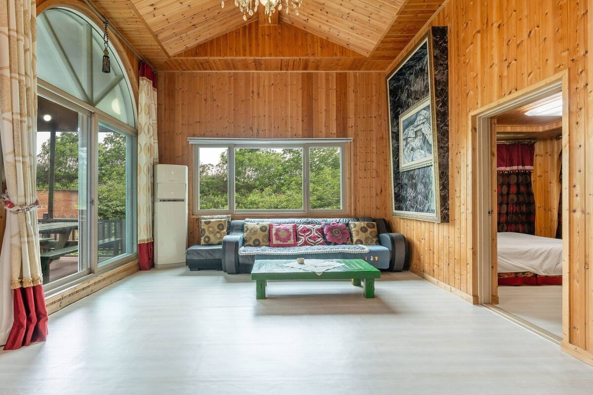 Une pièce spacieuse aux murs en bois et au plafond haut, dotée d’une grande fenêtre, d’un ensemble de canapés et d’une table basse verte.