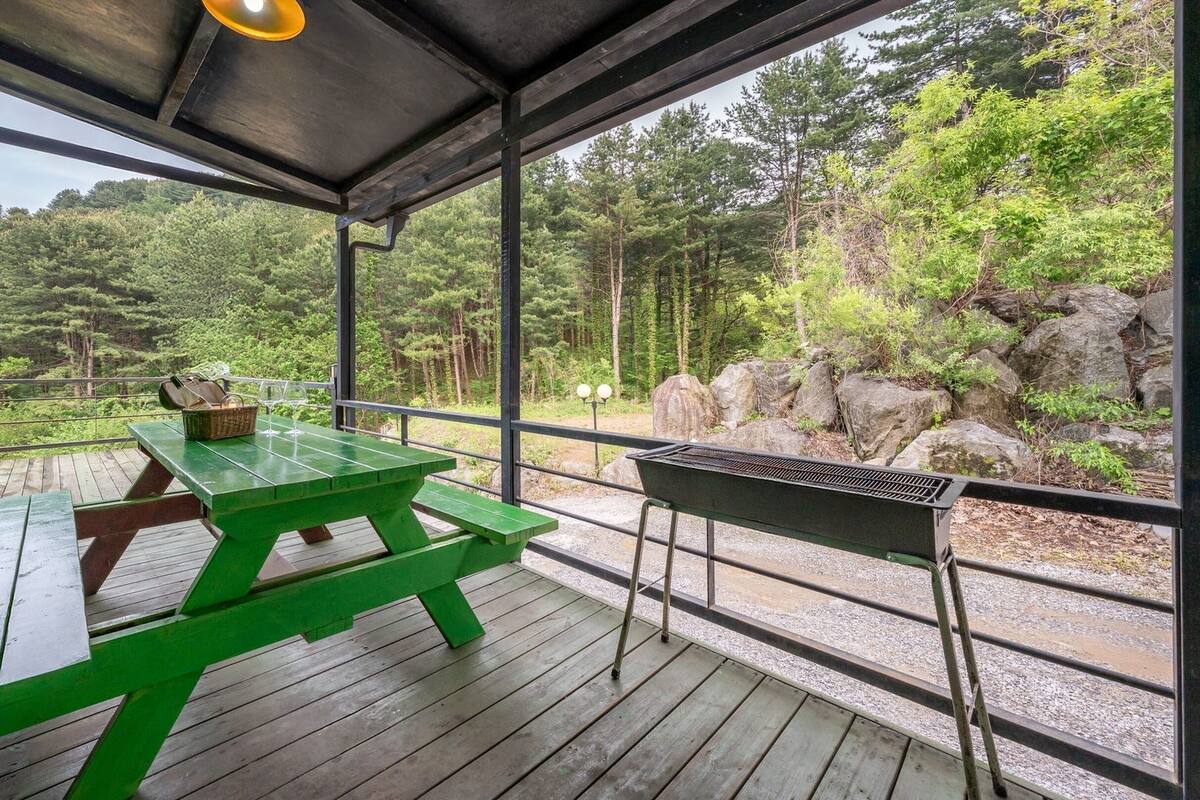 Une terrasse extérieure avec une table de pique-nique verte, un barbecue et une vue sur une zone boisée.