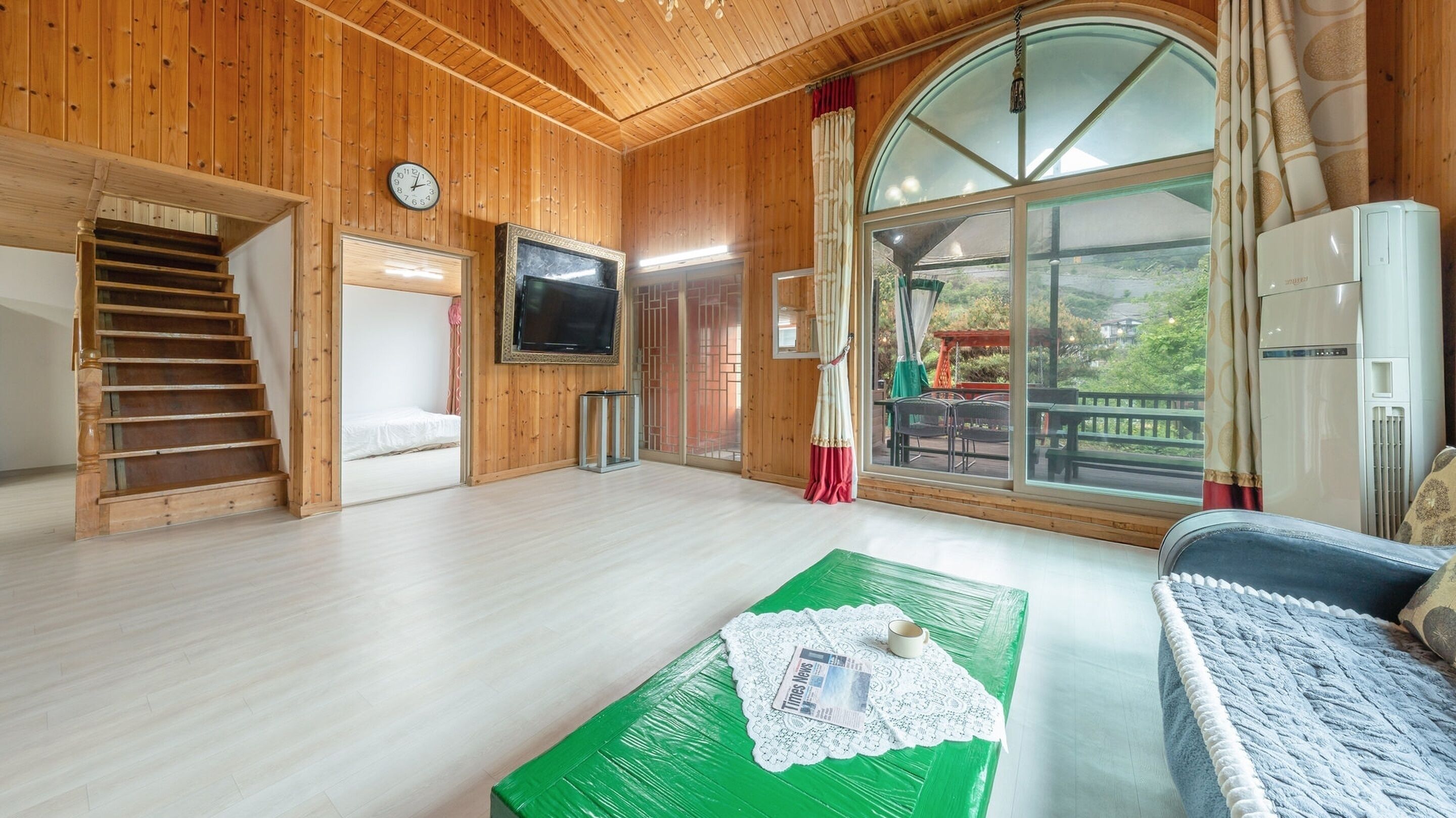 Un intérieur en bois avec un escalier, un téléviseur à écran plat, une table verte avec un journal et une grande fenêtre donnant sur un paysage.