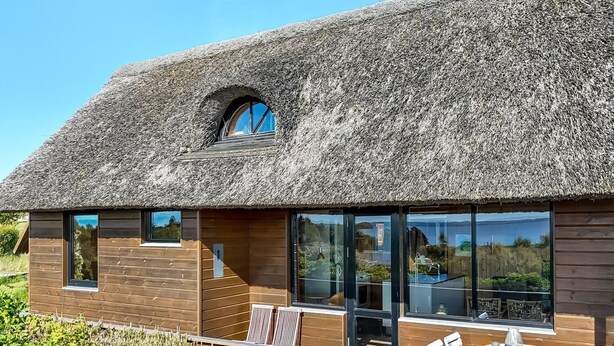 Une maison moderne avec un toit de chaume, de grandes fenêtres et un patio doté d’un espace salon extérieur.