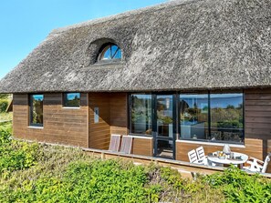 Ein modernes Haus mit Strohdach, großen Fenstern und einer Terrasse mit Sitzgelegenheiten.