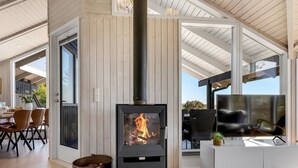 Una sala de estar moderna con una estufa de leña, un comedor con sillas y una mesa, y un televisor sobre un mueble blanco.
