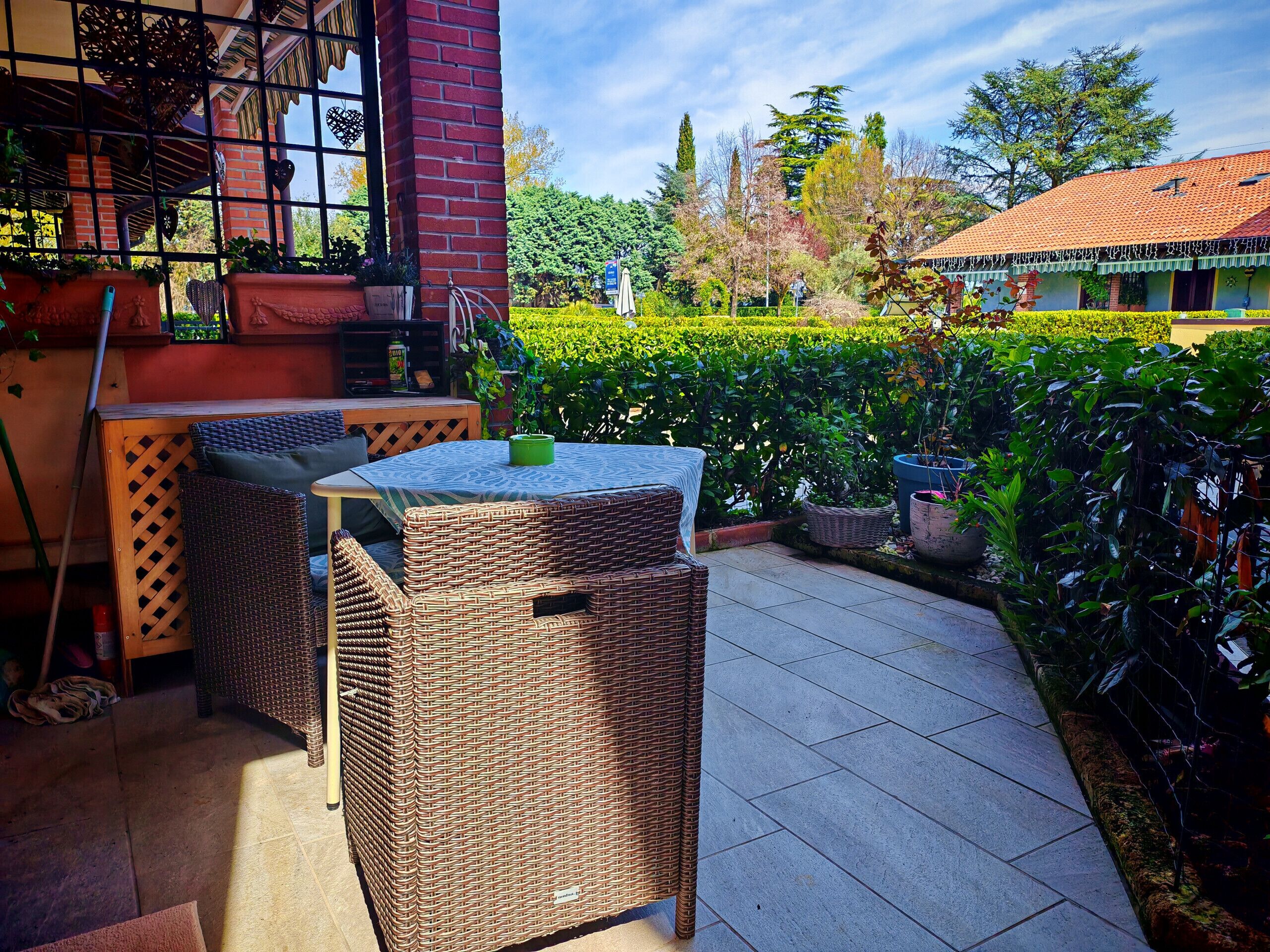 An outdoor patio with wicker furniture and a tiled floor, surrounded by greenery and a building with a red roof.