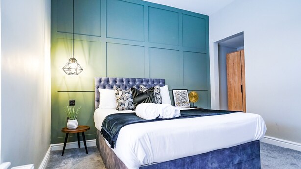 A bedroom with a tufted headboard, a bed with white linens, a small side table with a plant, and a hanging light fixture. - The City Centre Studios (Peterborough)