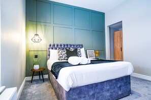 A bedroom with a tufted headboard, a bed with white linens, a small side table with a plant, and a hanging light fixture. - The City Centre Studios (Peterborough)