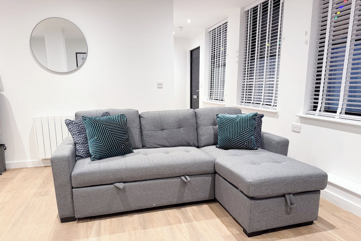 A grey sectional sofa with two patterned cushions, a round mirror, and a radiator in a modern room with white walls and wooden flooring.