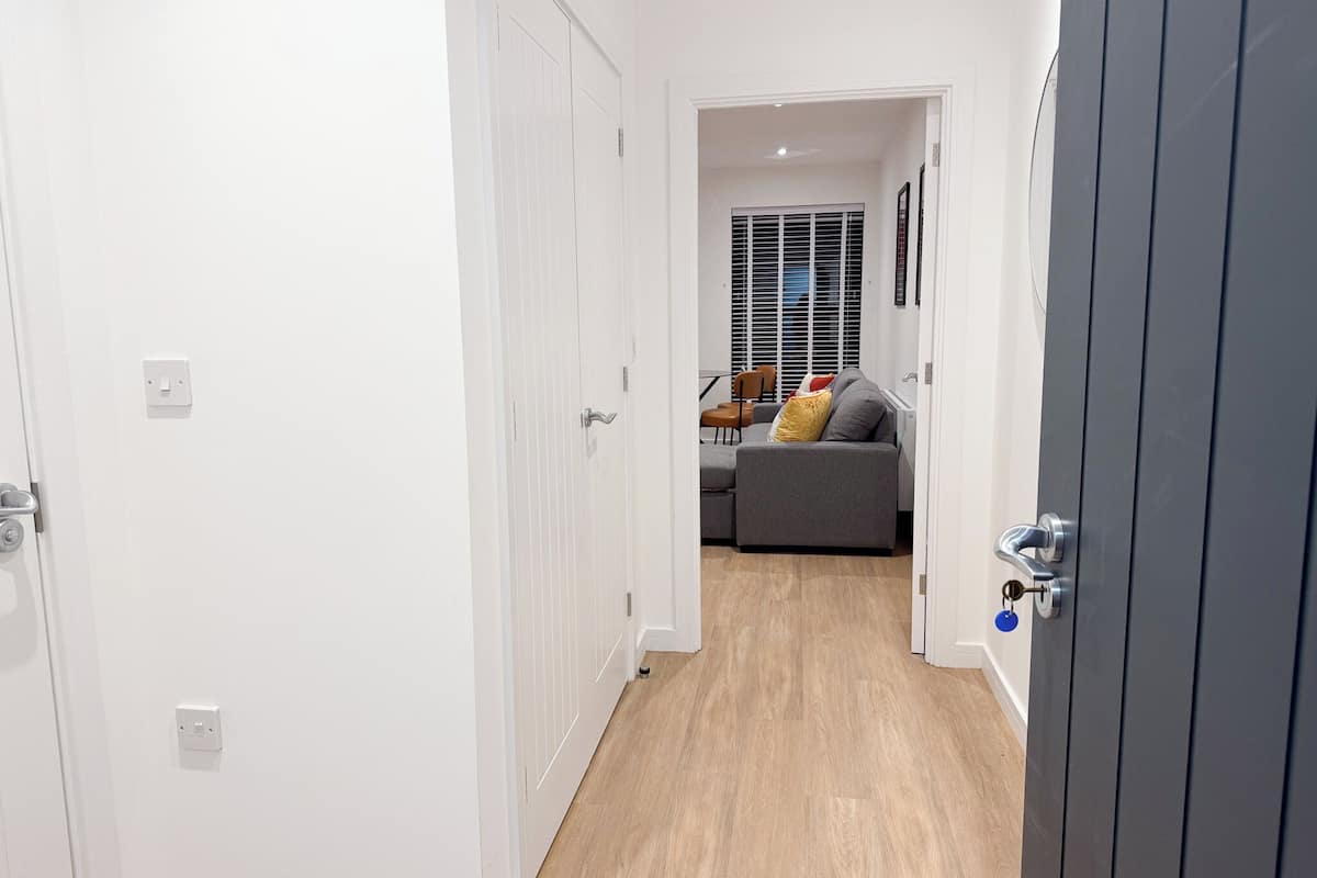 A narrow hallway with white walls and a wooden floor leading to a living area with a grey sofa and a window with blinds.