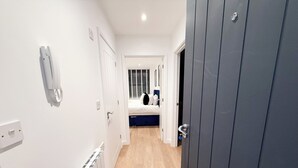 Un pasillo estrecho con un radiador, un teléfono y una puerta que da a un dormitorio con cama y ventana con persianas.