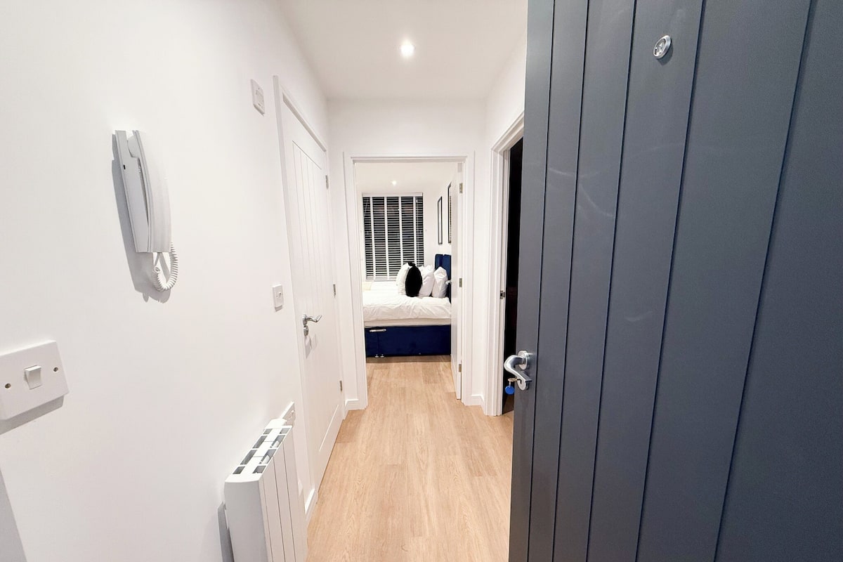 A narrow hallway with a radiator, a telephone, and a door leading to a bedroom with a bed and a window with blinds.