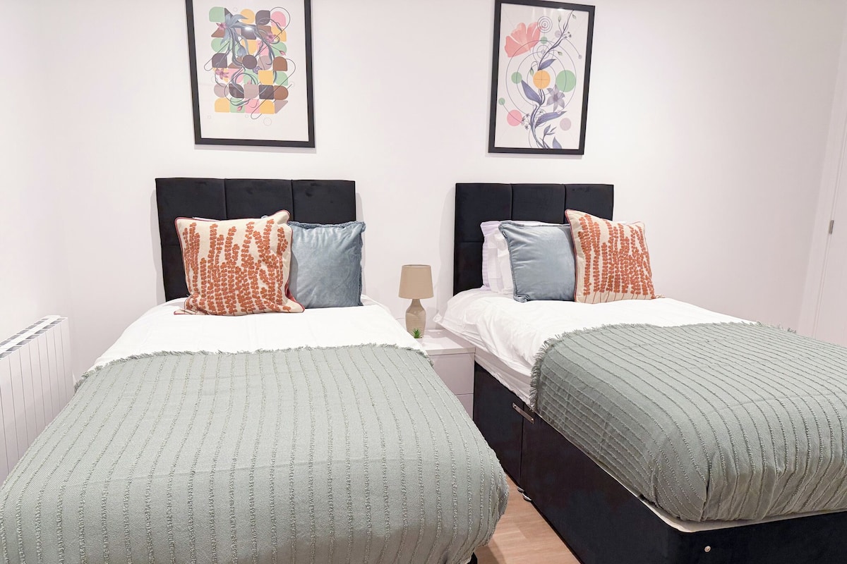 A twin room with two beds, each with a headboard, a bedside table, and a lamp. There are two framed floral artworks on the wall.