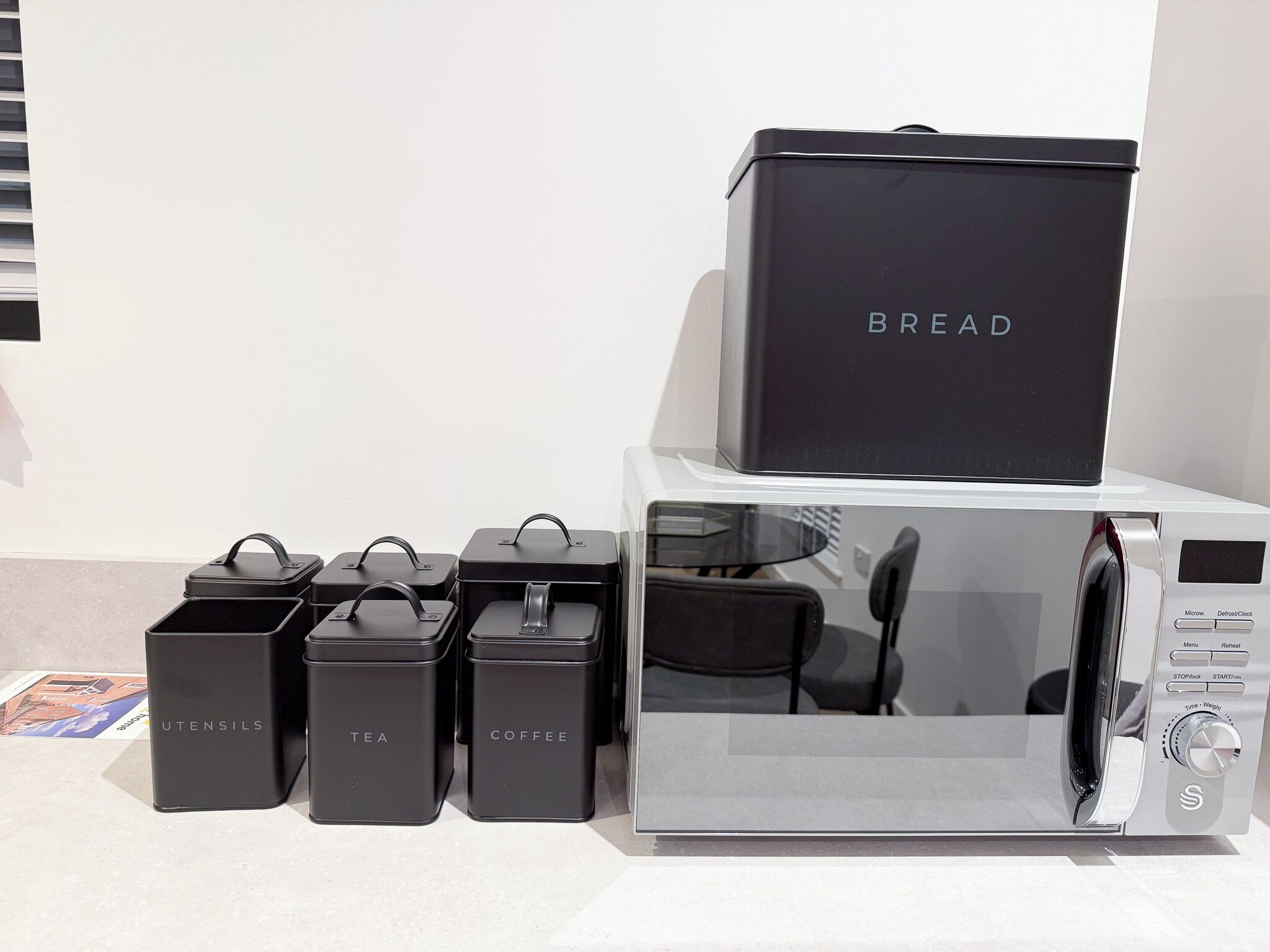 A kitchen with a microwave, a bread box, and three containers labeled "UTENSILS," "TEA," and "COFFEE."
