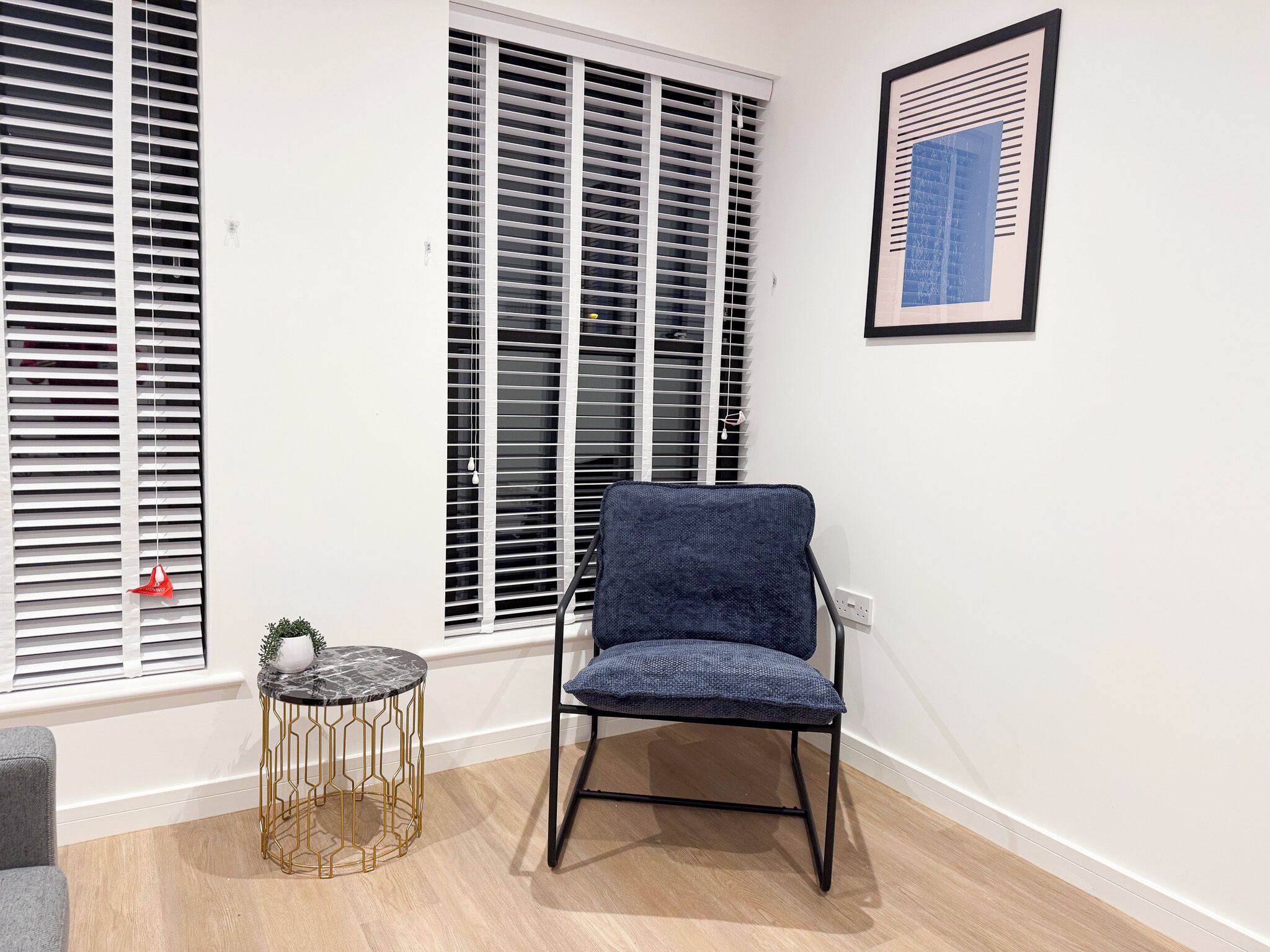A room with a blue chair, a small round table with a gold base, and a framed picture on the wall.