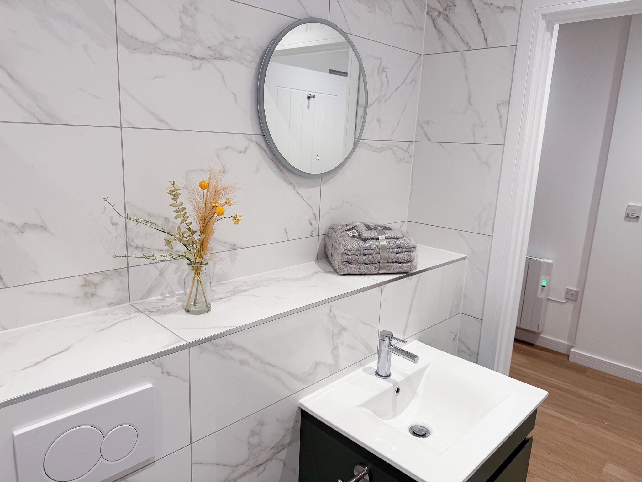 Un baño con lavamanos de mármol blanco, un espejo redondo y un florero con flores secas.