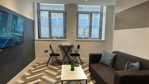 Una sala moderna con un sofá, una mesa de centro y dos sillones. Hay dos ventanas con persianas, un televisor de pantalla plana montado en la pared y un suelo con patrón de espiga.