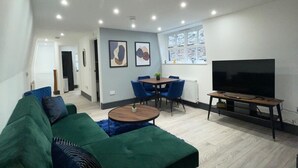 A modern living room with a green sofa, a round coffee table, a TV, and a dining area with blue chairs. - Abacus House Apartments (Huntingdon)