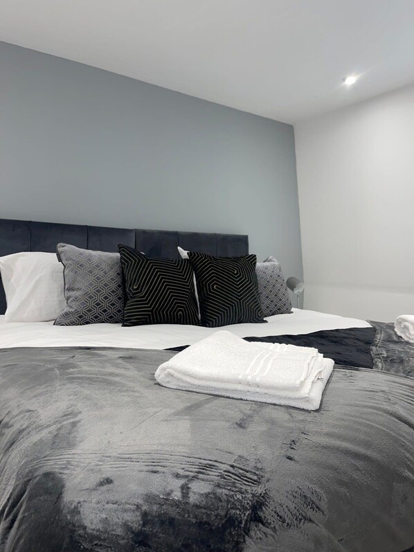 A modern bedroom with a grey headboard, a neatly made bed with patterned pillows, and a folded white towel on the bed. - Abacus House Apartments (Huntingdon)