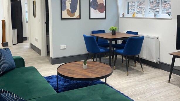 A modern living room with a green sofa, a round coffee table, and a dining area with blue chairs. There are two framed artworks on the wall and a window with a view of buildings. - Abacus House Apartments (Huntingdon)