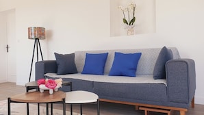 A modern living room with a grey sofa, blue cushions, a small round table with a vase of flowers, and a floor lamp.