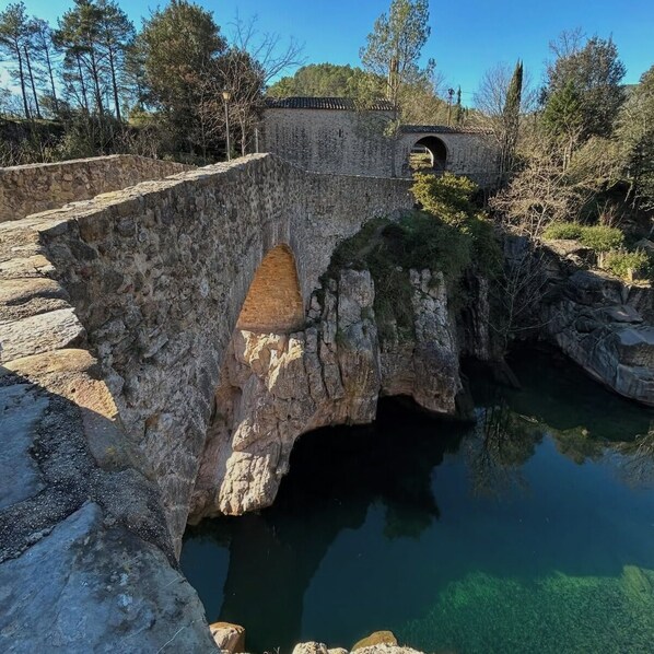 Un puente de piedra sobre un cuerpo de agua transparente, con un muro de piedra a un lado y árboles al fondo.