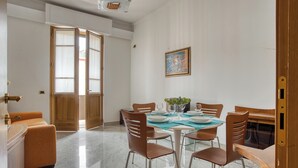 Zona pranzo con tavolo rotondo, sei sedie e piano in vetro. Una porta in legno dà accesso a un'altra stanza e c'è un piccolo divano arancione nell'angolo.