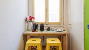 A small kitchenette with a window, two yellow stools, a coffee maker, a kettle, a vase with flowers, and a plate with snacks.