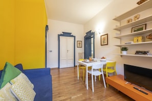 Ein Wohnzimmer mit blauem Sofa, einem Esstisch mit Stühlen, einem Fernseher und einem Bücherregal.