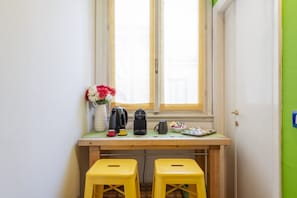 Eine kleine Küchenzeile mit Fenster, zwei gelbe Hocker, einer Kaffeemaschine, einem Wasserkocher, einer Vase mit Blumen und einem Teller mit Snacks.