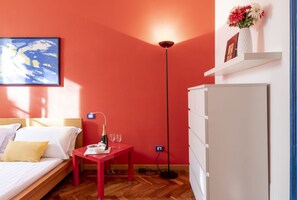 Ein Zimmer mit einem Bett, einem roten Tisch mit Flasche und Gläsern, einer Stehlampe und einem weißen Schrank.