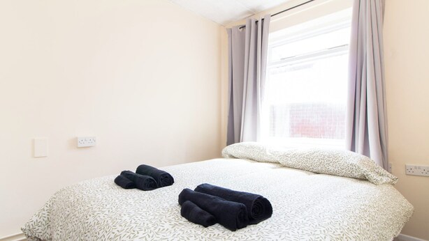 Een hotelkamer met een bed, twee opgerolde handdoeken, een raam met gordijnen en een stopcontact.