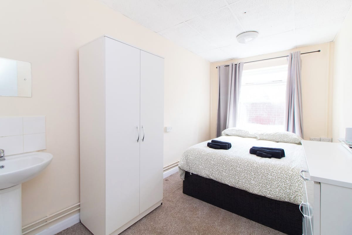 A bedroom with a bed, a wardrobe, a sink, and a window with curtains.