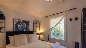 Una cama bien tendida con sábanas blancas y una toalla enrollada, una lámpara de noche, un cuadro en la pared, una ventana con cortinas y un radiador.