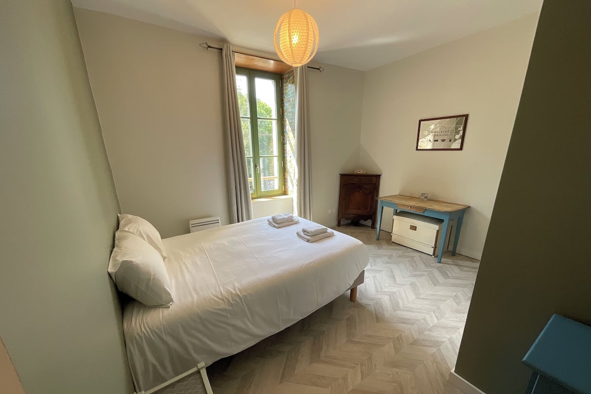 Un dormitorio con cama, escritorio, silla, ventana con cortinas y lámpara colgante.