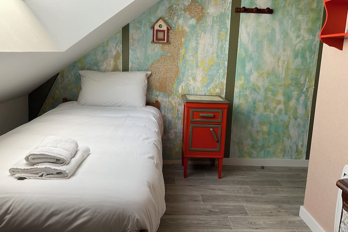 Un dormitorio con techo inclinado, una cama con ropa blanca, una mesita de noche roja y un comedero decorativo en la pared.
