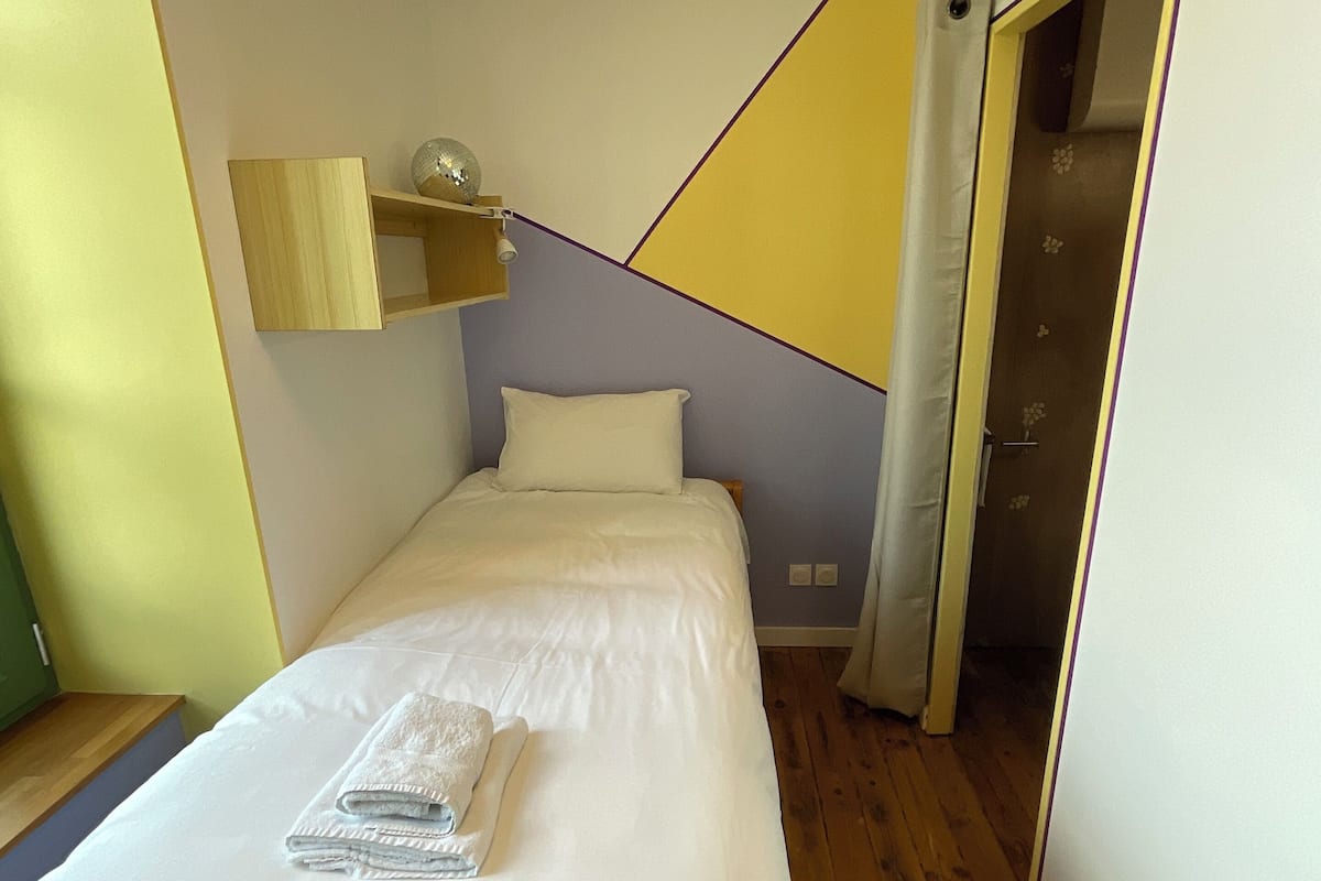 Un dormitorio con una cama, suelo de madera, estante en la pared y una puerta que da a otra habitación.