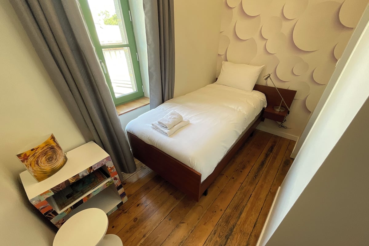 Un dormitorio ordenado con piso de madera, una cama con sábanas blancas, una mesita de noche con lámpara y una pequeña mesa redonda con un objeto decorativo.