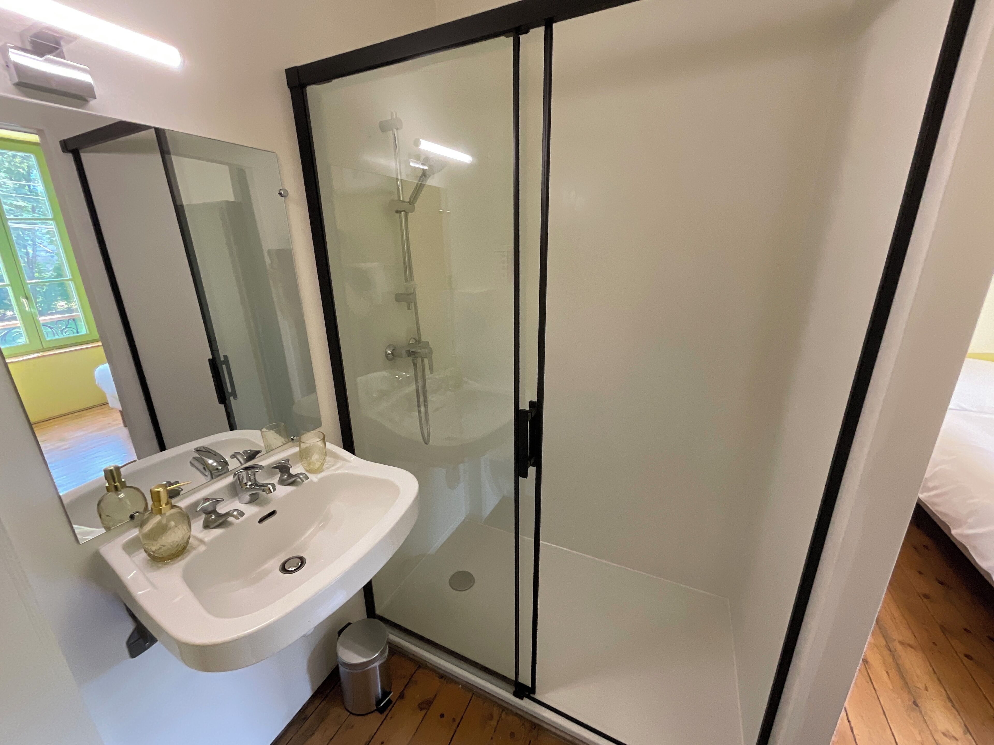 A modern bathroom with a glass-enclosed shower, a white sink with a mirror, and a small trash can.