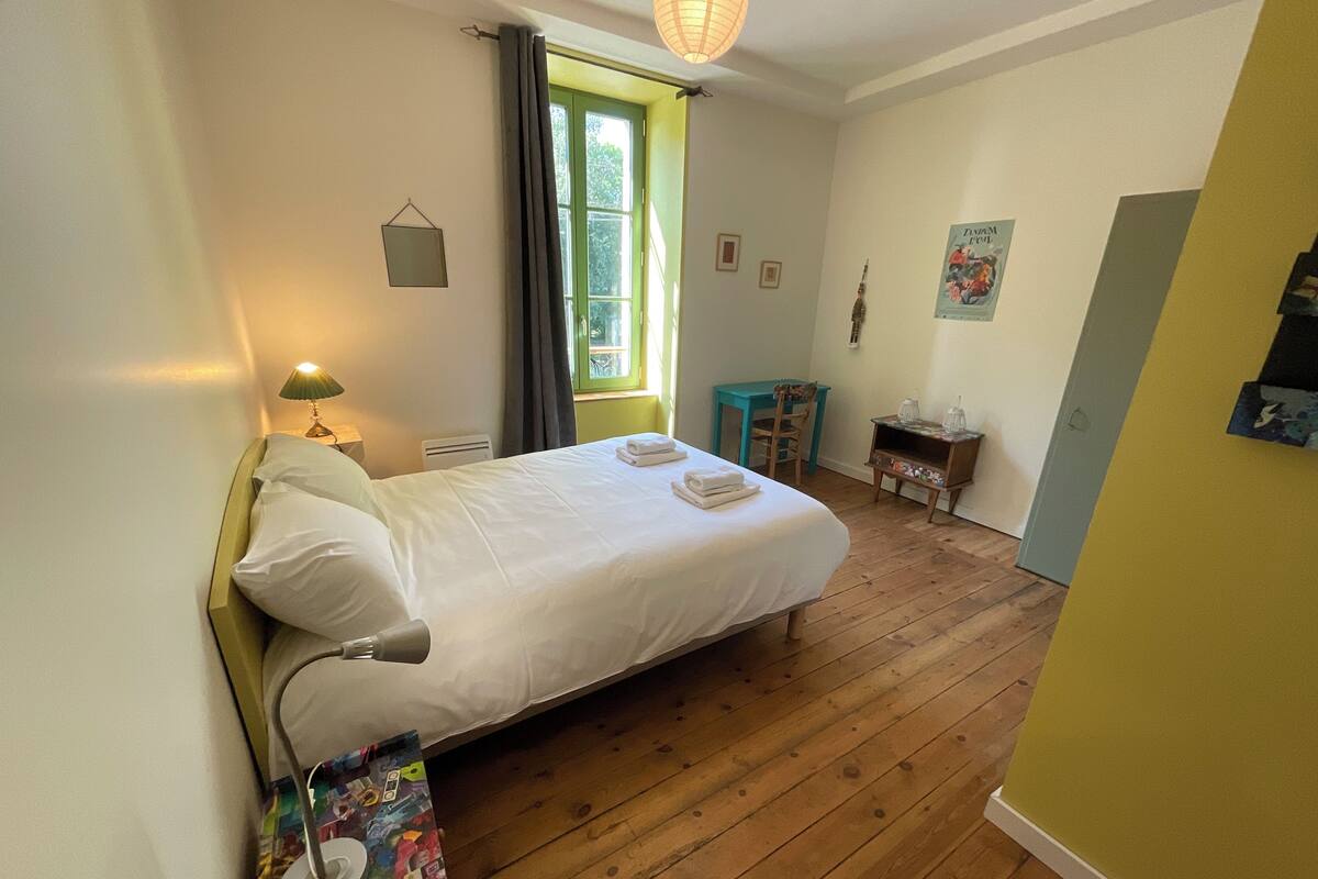 Un dormitorio con cama, lámpara de noche, suelo de madera, ventana con cortinas, una mesita con silla y una puerta.