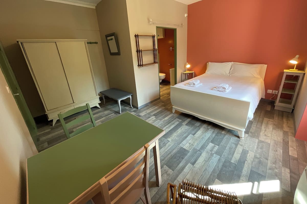Habitación de hotel con cama, escritorio, silla, armario y baño visible a través de una puerta abierta.