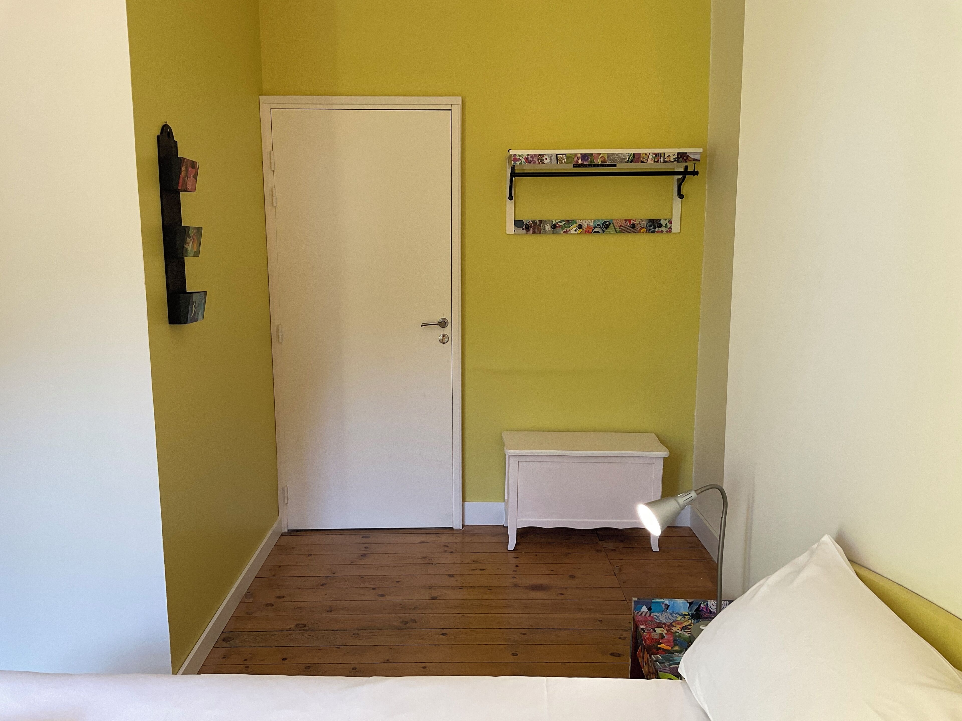 A small room with a wooden floor, a white door, a small white bedside table, a bedside lamp, and a colorful magazine rack.