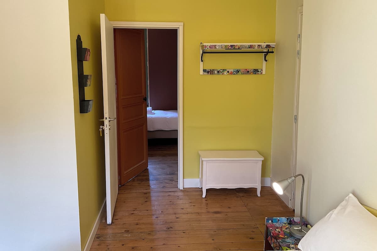 Un pasillo estrecho con piso de madera, una puerta a la izquierda, un pequeño banco blanco, un estante con objetos de colores y una cama al fondo.