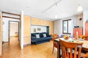 A room with a wooden floor, a blue sofa, a dining table with chairs, and a kitchen area with cabinets. - Casa Andy 1 Val Lavizzara With Views for Four (Lavizzara)