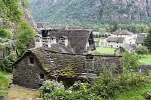 A stone building with a steep roof, surrounded by lush greenery and a mountainous backdrop. - Casa Andy 1 Val Lavizzara With Views for Four (Lavizzara)