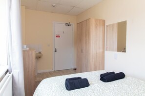 A hotel room with a bed, two towels, a wooden wardrobe, a bathroom with a sink, and a door with a sign.