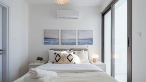 A modern bedroom with a large bed, a bedside table with a lamp, and a view of the sea through a large window. - Pauhad 3 - Larnaca Studios & Apts (Larnaca)